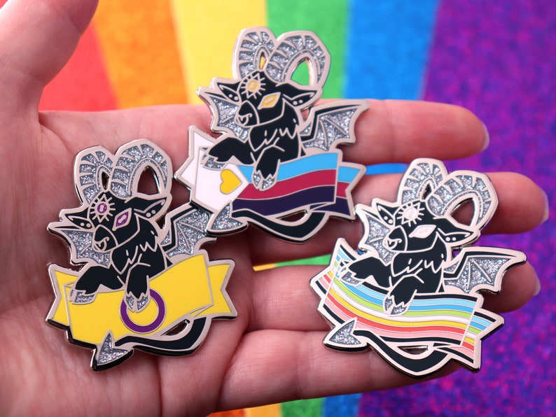 May include: Three black glitter enamel pins featuring a cartoon goat with horns and wings holding a banner. The banners feature different pride flags: the bisexual pride flag, the intersex pride flag, and the rainbow pride flag.
