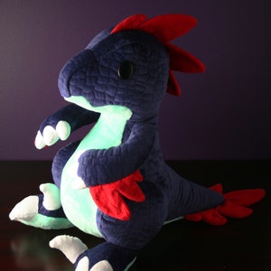 May include: A plush toy dinosaur with a blue body, red accents, and white claws. The dinosaur is sitting on a dark surface and has a glowing green light inside.