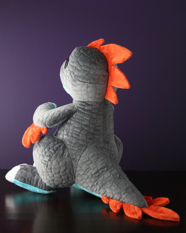 May include: A gray and turquoise plush dragon toy with orange spikes and tail. The dragon is sitting on a dark surface.