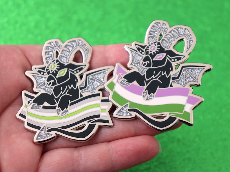 May include: Two silver glitter enamel pins featuring a black goat with wings and horns holding a green, white, and purple genderqueer pride flag.