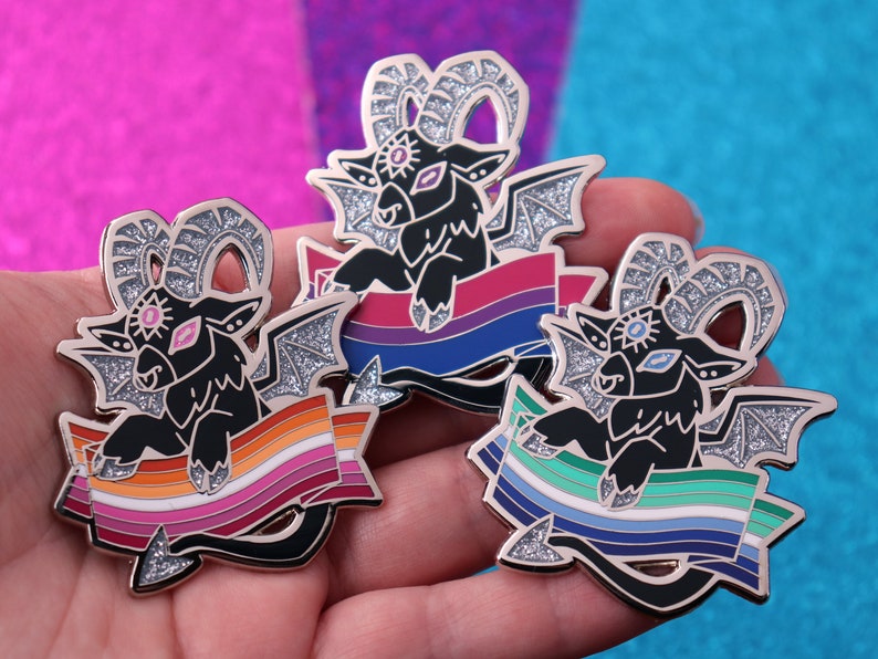 May include: Three enamel pins featuring a black goat with wings and a single eye, each holding a different pride flag. The flags are the lesbian pride flag, the bisexual pride flag, and the genderqueer pride flag.