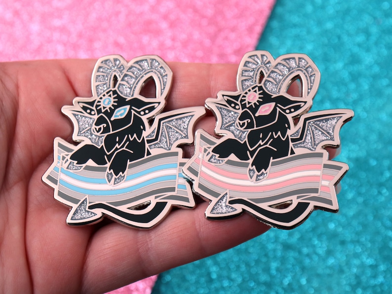 May include: Two gold-tone enamel pins featuring a black, winged goat with a third eye and horns. Each goat is holding a banner with a different color scheme: one with a grey, white, and light blue stripe pattern and the other with a pink, grey, and white stripe pattern.