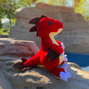 May include: A red plush dinosaur toy with black and brown accents and white claws. The dinosaur is sitting on a rock.