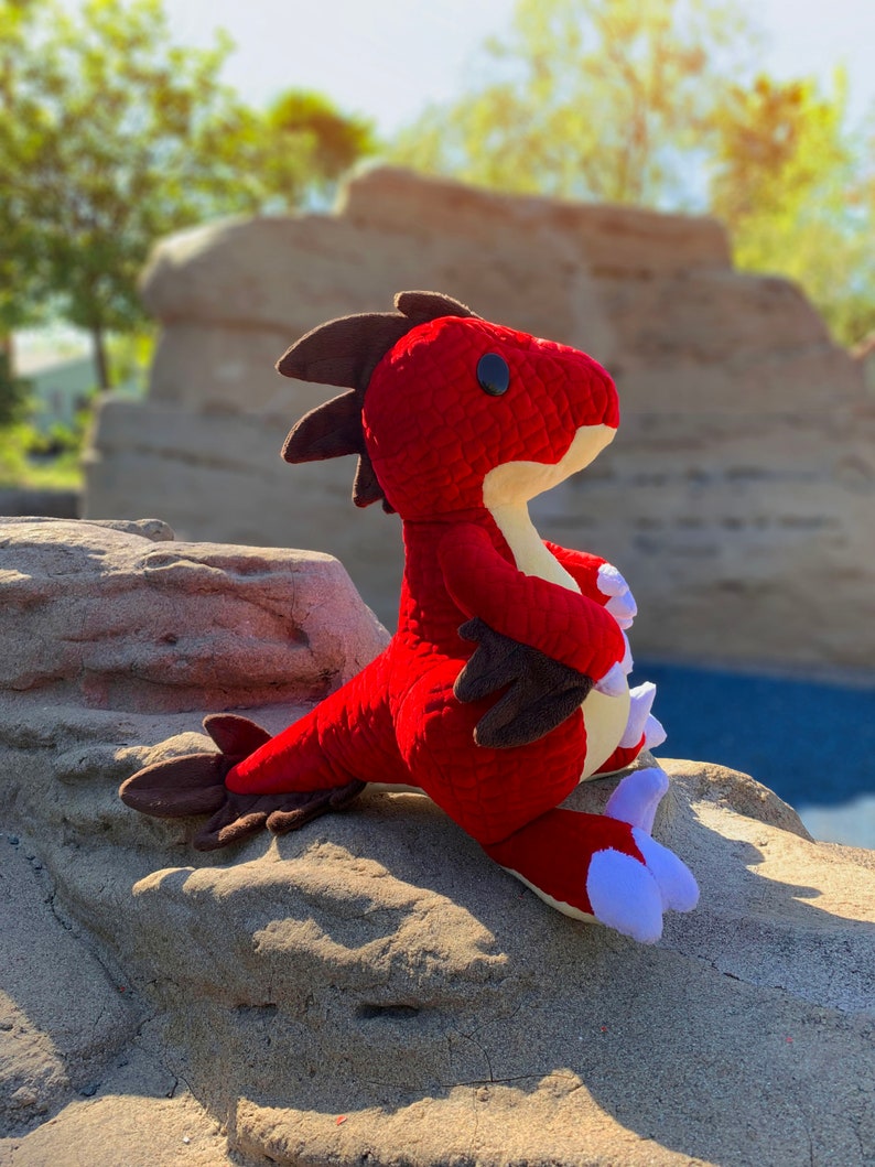 May include: A red plush dinosaur toy with black and brown accents and white claws. The dinosaur is sitting on a rock.