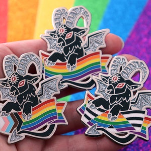 May include: Three silver enamel pins featuring a black goat with red eyes and horns, each holding a rainbow flag with different variations. The flags include a traditional rainbow, a transgender pride flag, and a bisexual pride flag.