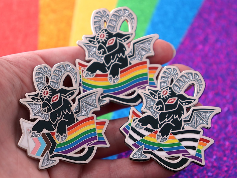 May include: Three silver enamel pins featuring a black goat with red eyes and horns, each holding a rainbow flag with different variations. The flags include a traditional rainbow, a transgender pride flag, and a bisexual pride flag.