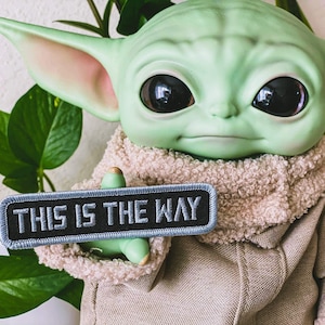 May include: A green plush toy of a character from the Star Wars franchise, holding a black and gray patch that reads "This is the way".