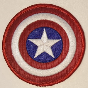 May include: A red, white, and blue embroidered patch featuring Captain America's shield. The shield is circular with a white star in the center on a blue background.