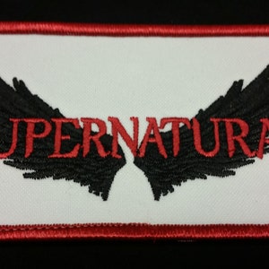 May include: Embroidered patch with the word "SUPERNATURAL" in red, arched over a pair of black wings. The patch has a white background and a red border.