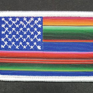 May include: A white embroidered patch with a blue field and white stars in the canton. The rest of the patch is a multi-colored striped design with red, orange, yellow, green, and blue stripes.