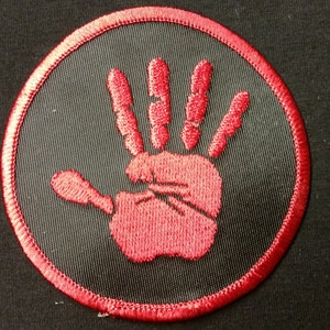 May include: Embroidered patch featuring a red handprint within a black circle, outlined with red stitching. The patch is set against a black fabric background, creating a striking contrast. Ideal for jackets, bags, or other apparel.