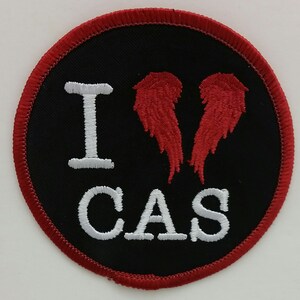 May include: Round black patch with the text "I CAS" in white, and red embroidered wings. The patch has a red border.