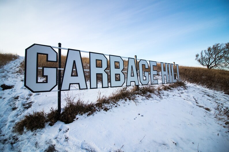 以下が含まれることがあります： 黒い文字で「GARBAGE HILL」と書いてある白い看板が、大きく太字で書かれています。看板は金属製のフェンスの支柱に取り付けられており、一部雪で覆われています。看板は、背景に小さな木がある雪の覆われた丘の上にあります。