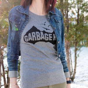 以下が含まれることがあります： 山脈と「GARBAGE ISLAND」という文字が黒で描かれたグレーのTシャツ。デニムジャケットを着た人が着ています。