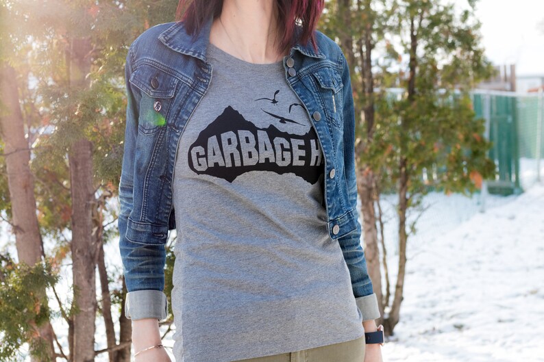 以下が含まれることがあります： 山脈と「GARBAGE ISLAND」という文字が黒で描かれたグレーのTシャツ。デニムジャケットを着た人が着ています。