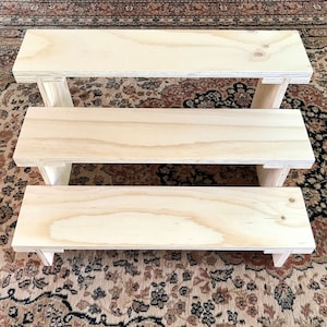 May include: A three-tiered, unfinished wooden display stand. The stand is constructed from light-coloured plywood, with each level supported by angled legs. The top tier measures approximately 61 cm long, the middle tier is 51 cm, and the bottom tier is 41 cm.