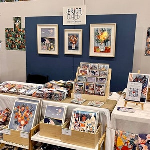 Puede incluir: Un puesto en una feria de artesan&iacute;a con una pared azul y un letrero que dice "Erica Walt Creative Studio". El puesto est&aacute; decorado con obras de arte enmarcadas, incluyendo estampados florales y paisajes. Tambi&eacute;n hay pilas de telas coloridas y tarjetas a la venta.