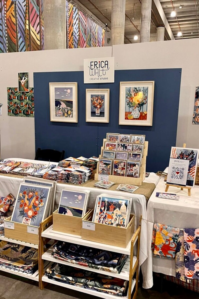 Puede incluir: Un puesto en una feria de artesan&iacute;a con una pared azul y un letrero que dice "Erica Walt Creative Studio". El puesto est&aacute; decorado con obras de arte enmarcadas, incluyendo estampados florales y paisajes. Tambi&eacute;n hay pilas de telas coloridas y tarjetas a la venta.