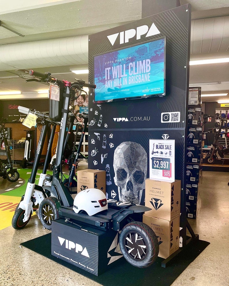 Puede incluir: Un scooter el&eacute;ctrico negro con un gr&aacute;fico de calavera y el texto "VIPPA PHANTOM" y "IT WILL CLIMB ANY HILL IN BRISBANE". El scooter tiene un casco blanco en la parte delantera y una caja negra con el texto "VIPPA" en el lateral. El scooter est&aacute; en exhibici&oacute;n en una tienda con un fondo blanco y negro.