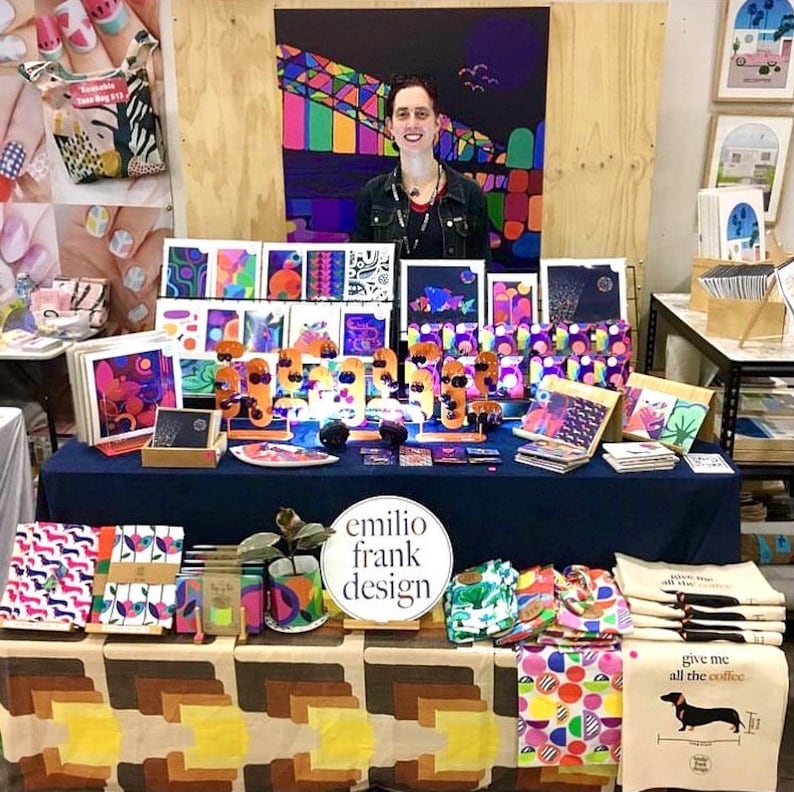 Puede incluir: Un puesto de mercado que muestra coloridas impresiones de arte abstracto, bolsas de tela y toallas de cocina. El puesto est&aacute; decorado con un letrero que dice "emilio frank design".