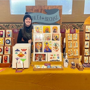Puede incluir: Un puesto de feria artesanal que presenta una variedad de estampados y tarjetas de arte coloridos. Las estampas presentan ilustraciones de mariposas, criaturas m&iacute;ticas y otros dise&ntilde;os caprichosos. La mesa est&aacute; decorada con un letrero de madera que dice "Hlea Bech&aacute;z".