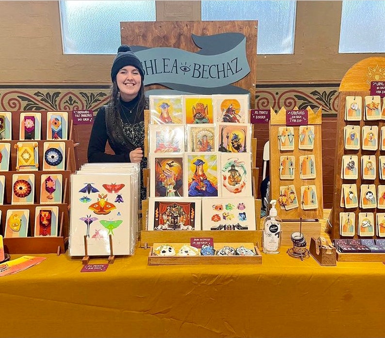 Puede incluir: Un puesto de feria artesanal que presenta una variedad de estampados y tarjetas de arte coloridos. Las estampas presentan ilustraciones de mariposas, criaturas m&iacute;ticas y otros dise&ntilde;os caprichosos. La mesa est&aacute; decorada con un letrero de madera que dice "Hlea Bech&aacute;z".