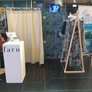 Puede incluir: Un soporte blanco con la palabra "farn" impresa en &eacute;l. El soporte est&aacute; delante de una cortina blanca. Un perchero de madera con un vestido estampado en azul y marr&oacute;n est&aacute; en el fondo.