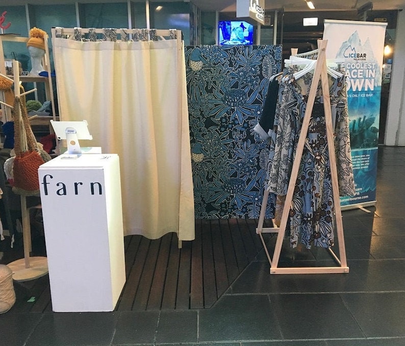 Puede incluir: Un soporte blanco con la palabra "farn" impresa en &eacute;l. El soporte est&aacute; delante de una cortina blanca. Un perchero de madera con un vestido estampado en azul y marr&oacute;n est&aacute; en el fondo.