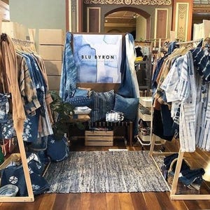 Puede incluir: Un escaparate de boutique de ropa que presenta una variedad de prendas te&ntilde;idas en &iacute;ndigo, incluyendo camisas, vestidos y bufandas. El escaparate est&aacute; instalado en estantes de madera e incluye un letrero que dice "BLU BYRON".