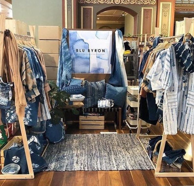 Puede incluir: Un escaparate de boutique de ropa que presenta una variedad de prendas te&ntilde;idas en &iacute;ndigo, incluyendo camisas, vestidos y bufandas. El escaparate est&aacute; instalado en estantes de madera e incluye un letrero que dice "BLU BYRON".