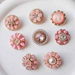 以下が含まれることがあります： さまざまな花柄や宝石のデザインの、8つの装飾ボタンのコレクション。ボタンはピンク、ゴールド、パールをアクセントとしています。ラインストーンやパールの装飾が施されたものもあれば、滑らかで光沢のある仕上がりのものもあります。白い表面に配置されています。
