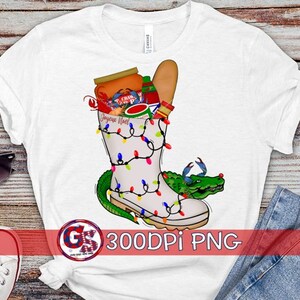 May include: White t-shirt with a graphic of a boot filled with Christmas lights, a crawfish, and other holiday items. The boot is surrounded by a green alligator. The text "Joyeux Noel" is on the boot.