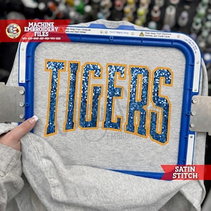 May include: A light gray sweatshirt fabric secured in a blue plastic embroidery hoop. The word TIGERS is embroidered in the center with blue glittery thread and a thick orange satin stitch outline, creating a bold, textured collegiate design.