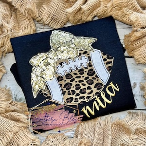 Puede incluir: Una camiseta negra con un lazo de lentejuelas doradas y una pelota de fútbol con un diseño de estampado de leopardo. La pelota de fútbol tiene cordones blancos y un contorno blanco. El nombre "mia" está bordado en dorado debajo de la pelota de fútbol.