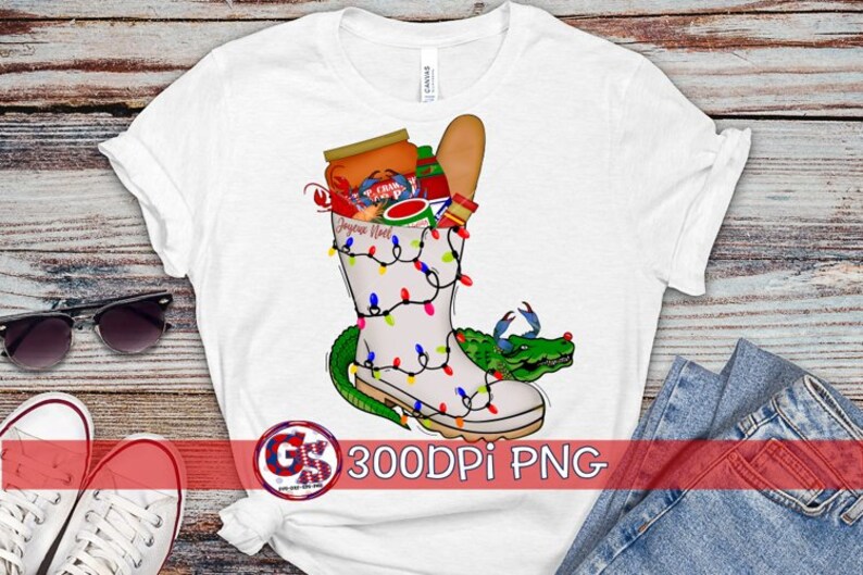 May include: White t-shirt with a graphic of a boot filled with Christmas lights, a crawfish, and other holiday items. The boot is surrounded by a green alligator. The text "Joyeux Noel" is on the boot.