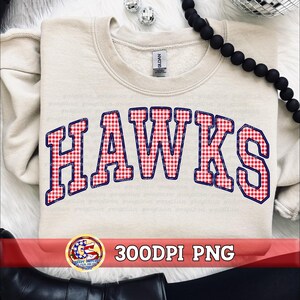 Könnte beinhalten: Ein beigefarbenes Sweatshirt mit Rundhalsausschnitt und dem Wort HAWKS in einem rot-weißen Vichy-Karomuster mit dunkelblauem Rand. Der Schriftzug ist bogenförmig auf der Brust platziert. Das Kleidungsstück hat ein Gildan-Etikett am Innenkragen.