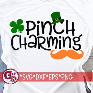 May include: White t-shirt with a St. Patrick's Day design. The design includes a green shamrock, a green leprechaun hat, an orange mustache, and the words "Pinch Charming" in black. The bottom of the shirt has a logo and file type information.