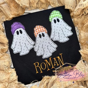 May include: Black fabric featuring three ghost appliqués, each with a different patterned hat in purple, orange, and green. The name "ROMAN" is embroidered in orange. The design is framed by burlap.