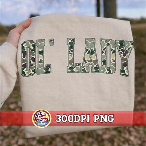 May include: A beige sweatshirt featuring the text "OL' LADY" in a block font. The letters are filled with a green, brown, and tan camouflage pattern and outlined in a dark green border. The fabric is a solid light neutral color throughout.