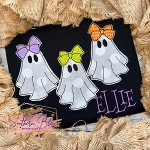 May include: Black fabric featuring three ghost designs, each with a different coloured bow. The ghosts are white with grey polka dots. The name "ELLIE" is embroidered in purple. The logo "Southern Bleu Embroidery" is in the lower left corner.