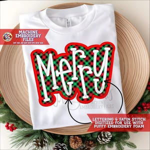Puede incluir: Camiseta blanca con la palabra "Merry" en un diseño de cuadros rojos y verdes. Las letras son blancas con un contorno rojo. La camiseta se muestra en una bandeja de madera con decoraciones navideñas.