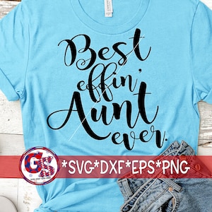 May include: A light blue t-shirt with the phrase "Best effin' Aunt ever" in black script. The shirt is folded, with a pair of red sunglasses and denim shorts visible. A logo with text is at the bottom.