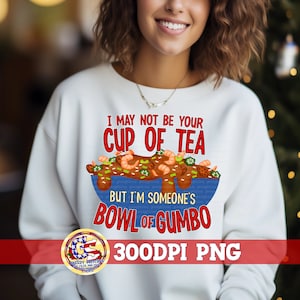 May include: White sweatshirt with a red and blue graphic design. The graphic features a bowl of gumbo with the text "I may not be your cup of tea, but I'm someone's bowl of gumbo".