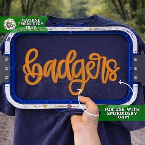 May include: A dark blue t-shirt secured in a rectangular blue embroidery hoop. The fabric features the word "badgers" stitched in a thick, raised orange thread. The hoop includes white plastic edges with metal screws and printed text markings.