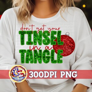 May include: White sweatshirt with the text "don't get your TINSEL in a TANGLE" in green and red. A red glittery Christmas ornament graphic is on the right. The bottom of the image has a logo and the text "300DPI PNG".
