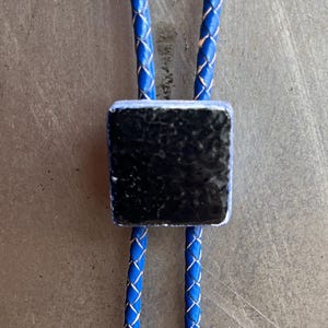 May include: A blue bolo tie with a square, textured black centerpiece. The cord is a vibrant blue with a white accent. This Western-style accessory is a classic piece of jewelry.