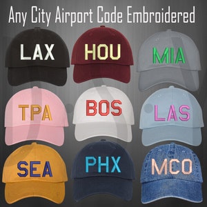May include: Nine embroidered baseball caps with different city airport codes. The caps are in various colors, including black, red, gray, pink, white, blue, yellow, navy, and denim.