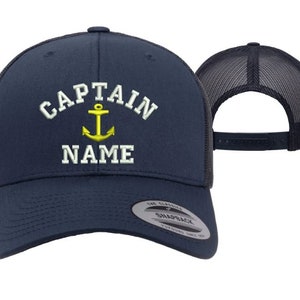 May include: A navy blue trucker hat with a white embroidered anchor and the text "CAPTAIN NAME". The hat has a mesh back and a snap closure.