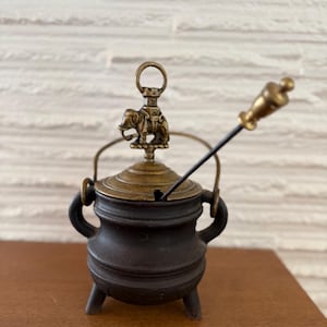 May include: A small black cast iron pot with a brass lid and handle. The pot has three legs and a brass elephant figurine on top of the lid. A small brass rod with a knob on the end is attached to the pot.