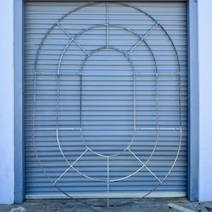 May include: A large, metal, oval-shaped sculpture composed of interconnected curved and straight bars. The sculpture is mounted on a gray, corrugated metal surface, framed by a light blue border and white wall. A yellow measuring tape lies on the ground.